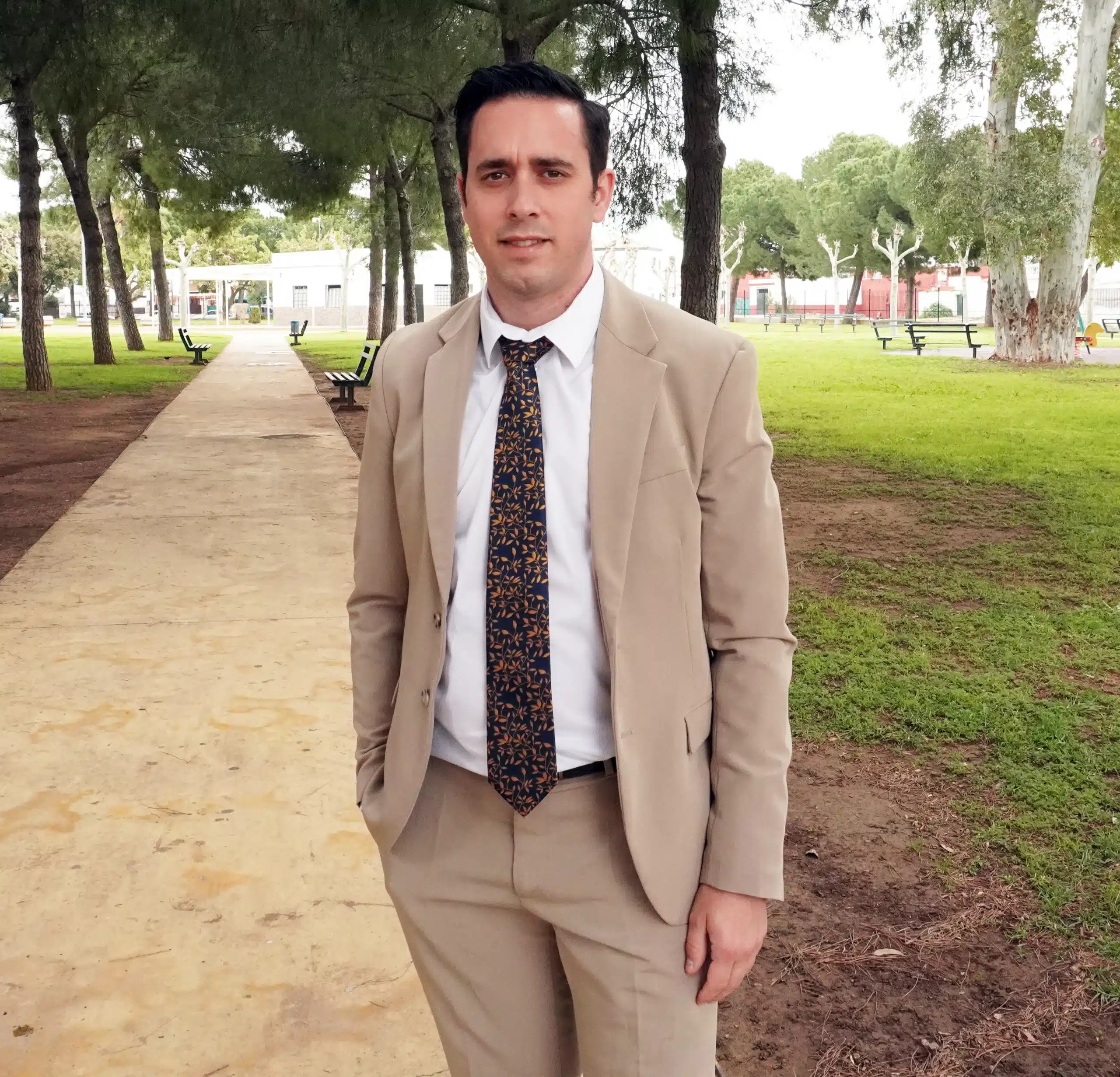 Nuestro agente Adrian, vestido con un traje de color beige y una corbata estampada, posa para la cámara en un parque.