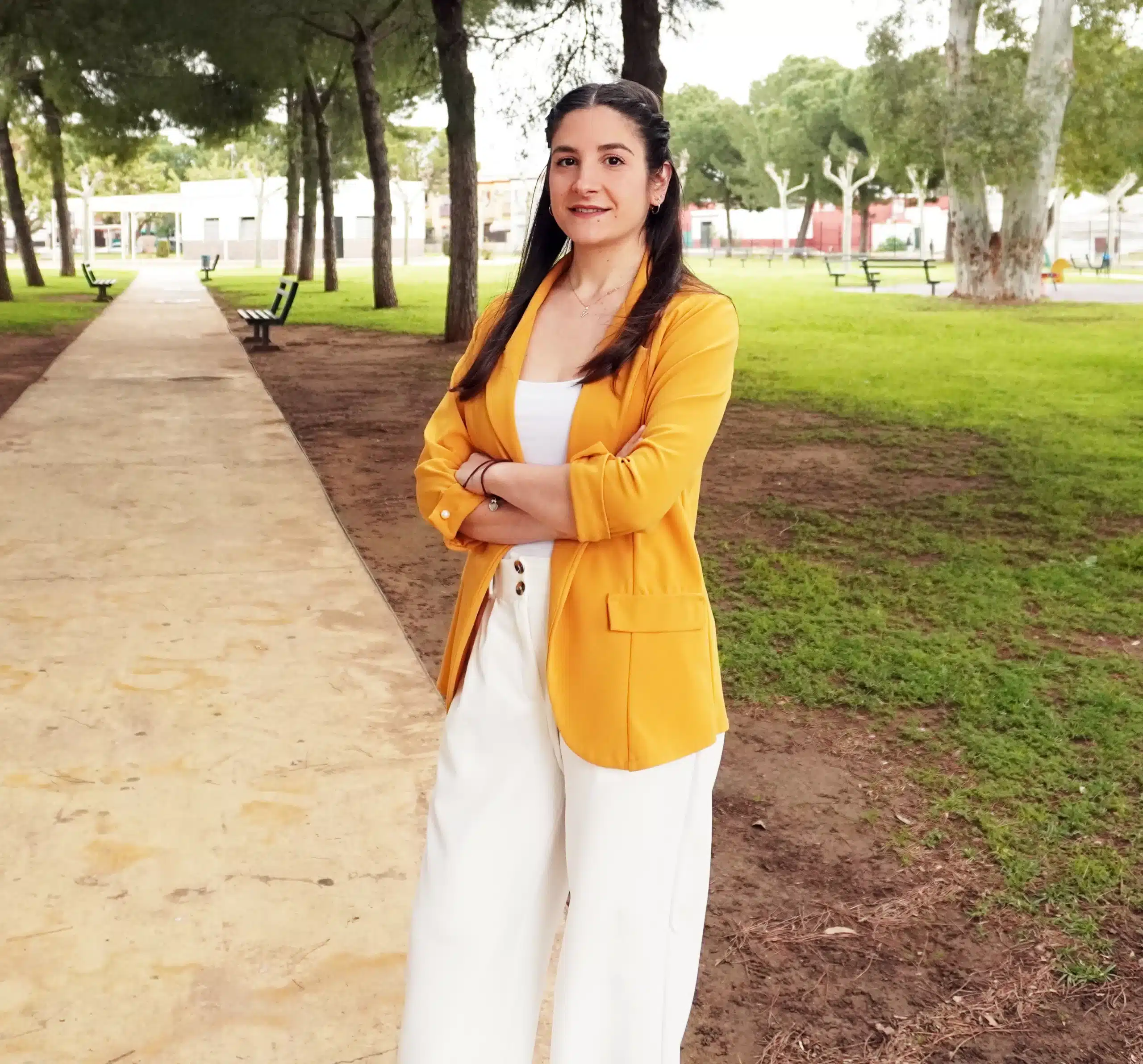 Nuestra agente Macarena, vestida con una chaqueta amarilla, pantalón blanco y blusa blanca, sonríe y posa con los brazos cruzados en un parque.