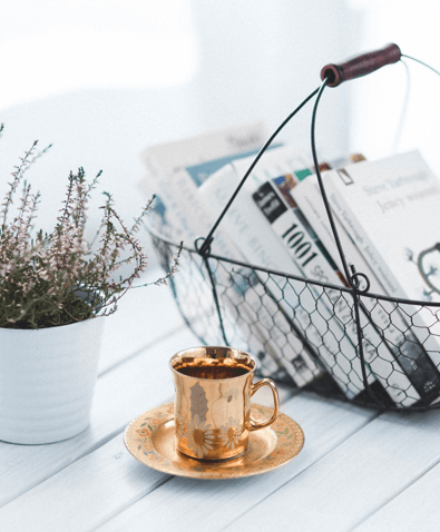 Una cesta de metal con libros, una taza de café dorada y una planta de interior sobre una mesa de madera blanca.