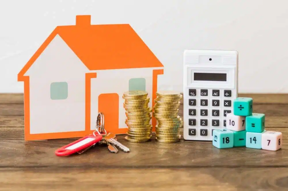 Cálculo y Planificación de la Hipoteca Una maqueta de una casa de papel, junto a la cual hay unas llaves, pilas de monedas, una calculadora y unos cubos de colores con números.