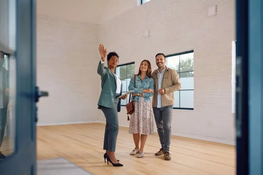 Una agente inmobiliaria, de raza negra y vestida con un traje verde, le muestra un espacio vació a una pareja caucásica, señalando con la mano.