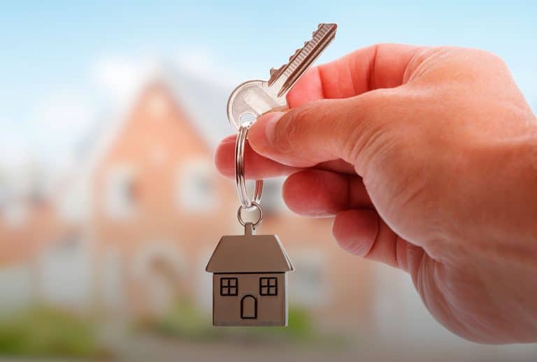 Una mano sostiene unas llaves con un llavero en forma de casa, con una casa sifuminada de fondo.