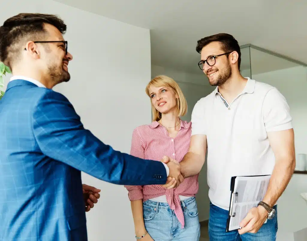 Un agente inmobiliario, vestido con un traje azul, estrecha la mano de un cliente. La pareja del cliente sonríe y los mira.