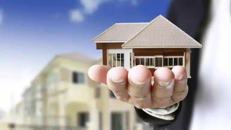 Una persona, vestida con un traje oscuro, sostiene en la palma de su mano una pequeña maqueta de una casa, con otra casa real y un cielo azul en el fondo. Servicios inmobiliarios