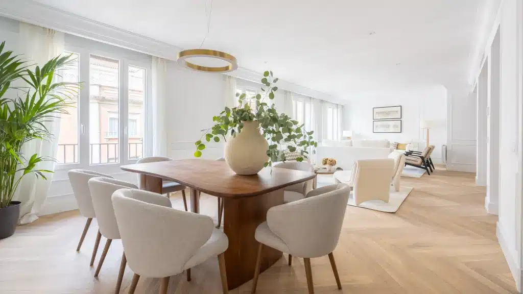 Un amplio salon comedor, decorado con un estilo moderno y minimalista, con mesa de madera, sillas claras, un gran jarrón con ramas y un luminoso salón al fondo