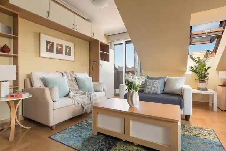 Una sala de estar en un ático, con techos inclinados y una ventana de techo, que muestra un sofá de dos plazas, un sofá cama, una mesa de café y estantes.