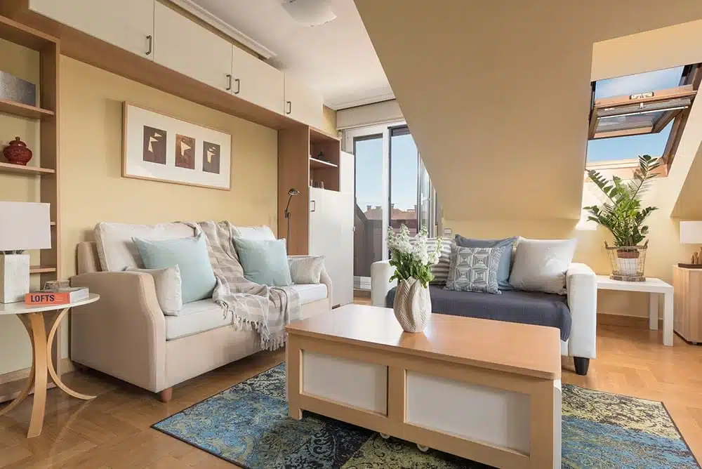 Una sala de estar en un ático, con techos inclinados y una ventana de techo, que muestra un sofá de dos plazas, un sofá cama, una mesa de café y estantes.