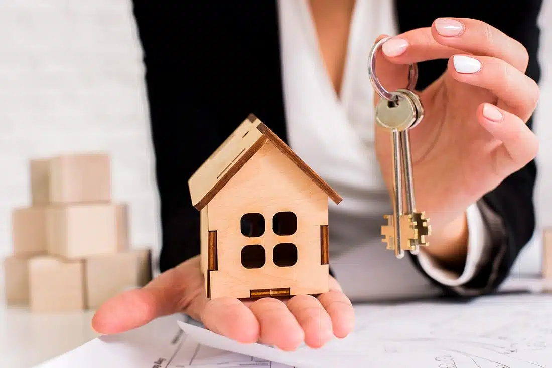 Una persona, presumiblemente una agente inmobiliaria, sostiene en una mano una maqueta de una casa de madera y en la otra, las llaves de la casa. En el fondo se ven unas cajas de madera.