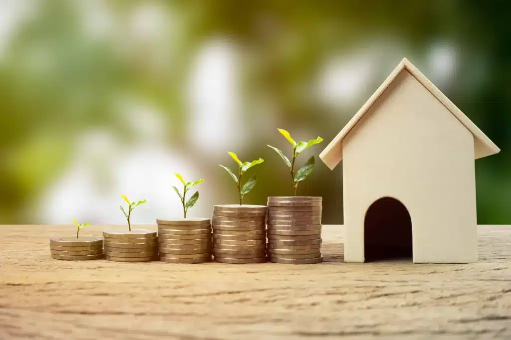 Una maqueta de una casa de madera junto a una serie de pilas de monedas de altura creciente, con pequeñas plantas germinando de cada una de ellas, simbolizando el crecimiento de la inversión.