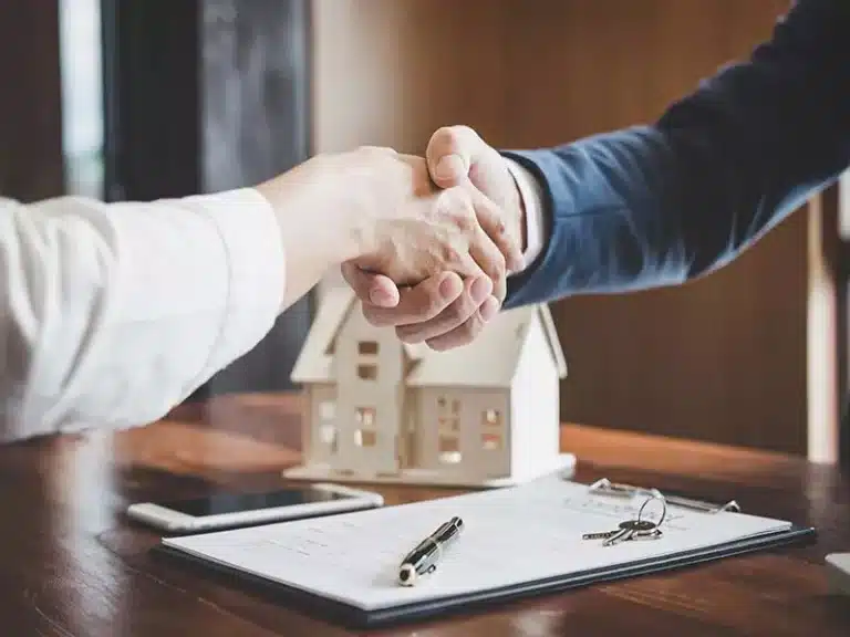 Dos personas dándose la mano sobre un escritorio con documentos y una maqueta de una casa.