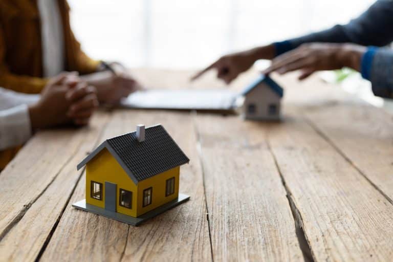 Maqueta de casa amarilla en primer plano sobre una mesa de madera. En el fondo, dos personas interactúan con documentos, una señalando un punto y la otra con un bolígrafo, en la firma de un contrato.