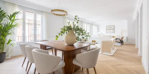 Un amplio salon comedor, decorado con un estilo moderno y minimalista, con mesa de madera, sillas claras, un gran jarrón con ramas y un luminoso salón al fondo